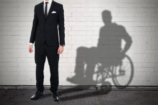 A man in standing in front of a white wall, his shadow behind him shows him sitting down in a wheelchair.