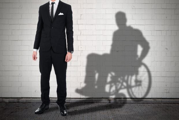 A man is standing, his shadow behind him shows him sitting in a wheelchair.