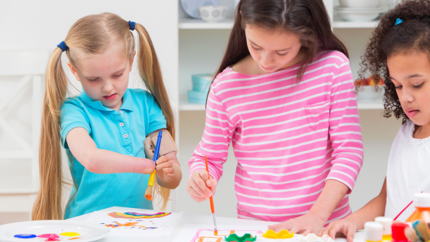 Three young girls paint together at a table, one has a visible disability.