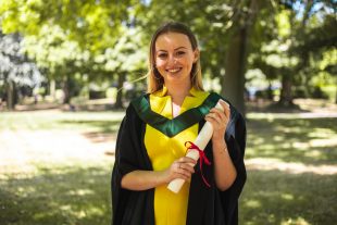 Eleanor is standing outside at her graduation, wearing a yellow dress and dark green and black graduation robes. She is holding a degree scroll and smiling.