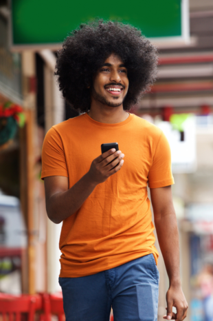 A black man smiles as he walks down the street, he has his phone held up in front of him as if he is texting.