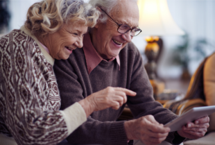 An elderly man and woman smile while looking at a tablet.