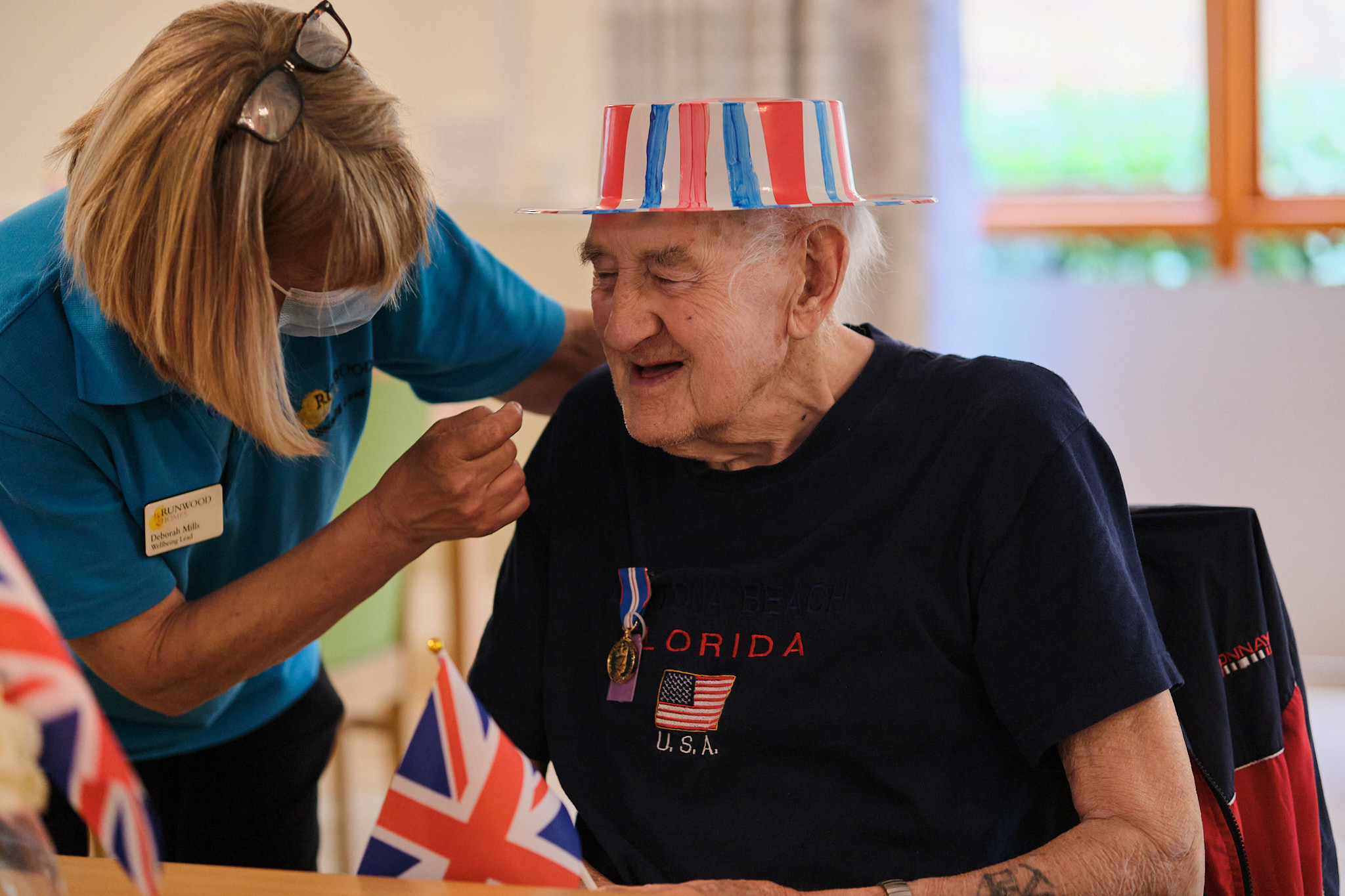 A smiling older gentleman in a Union Jack hat and dark blue shirt holds a Union Jack flag whilst a care home worker in a mask leans over to talk to him. 
