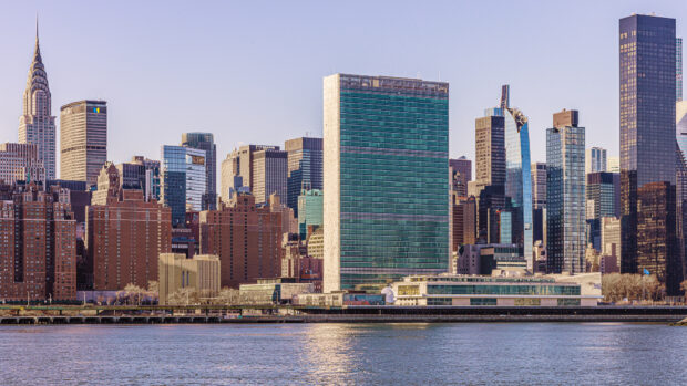 Image of New York skyline in front of the river, with the UN Headquarters building which has green glazing in the middle of the scene.