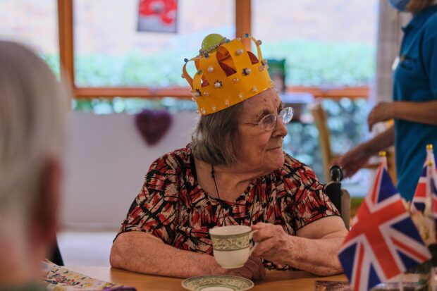 Lady sat down holding a cup and wearing a paper made crown decorated in beads and gems. 