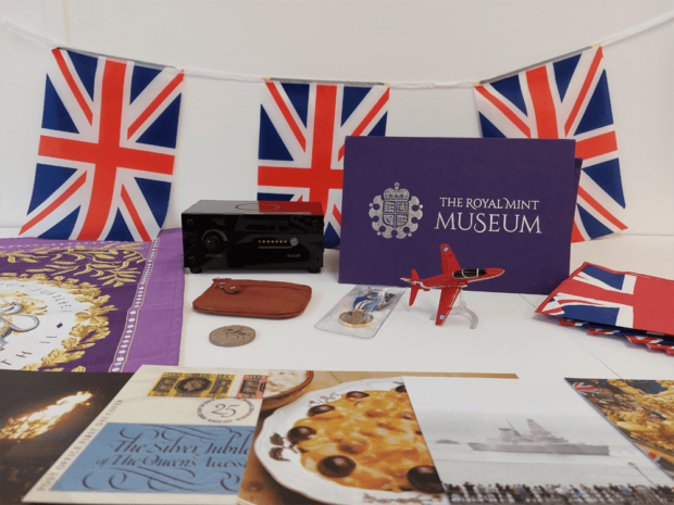 The contents of a 'Museum in a box' with Union Jack bunting and flags, a Jubilee teatowel, recipes from the Silver Jubilee, a picture of a warship, a model airplane, a small red purse and a coin. 