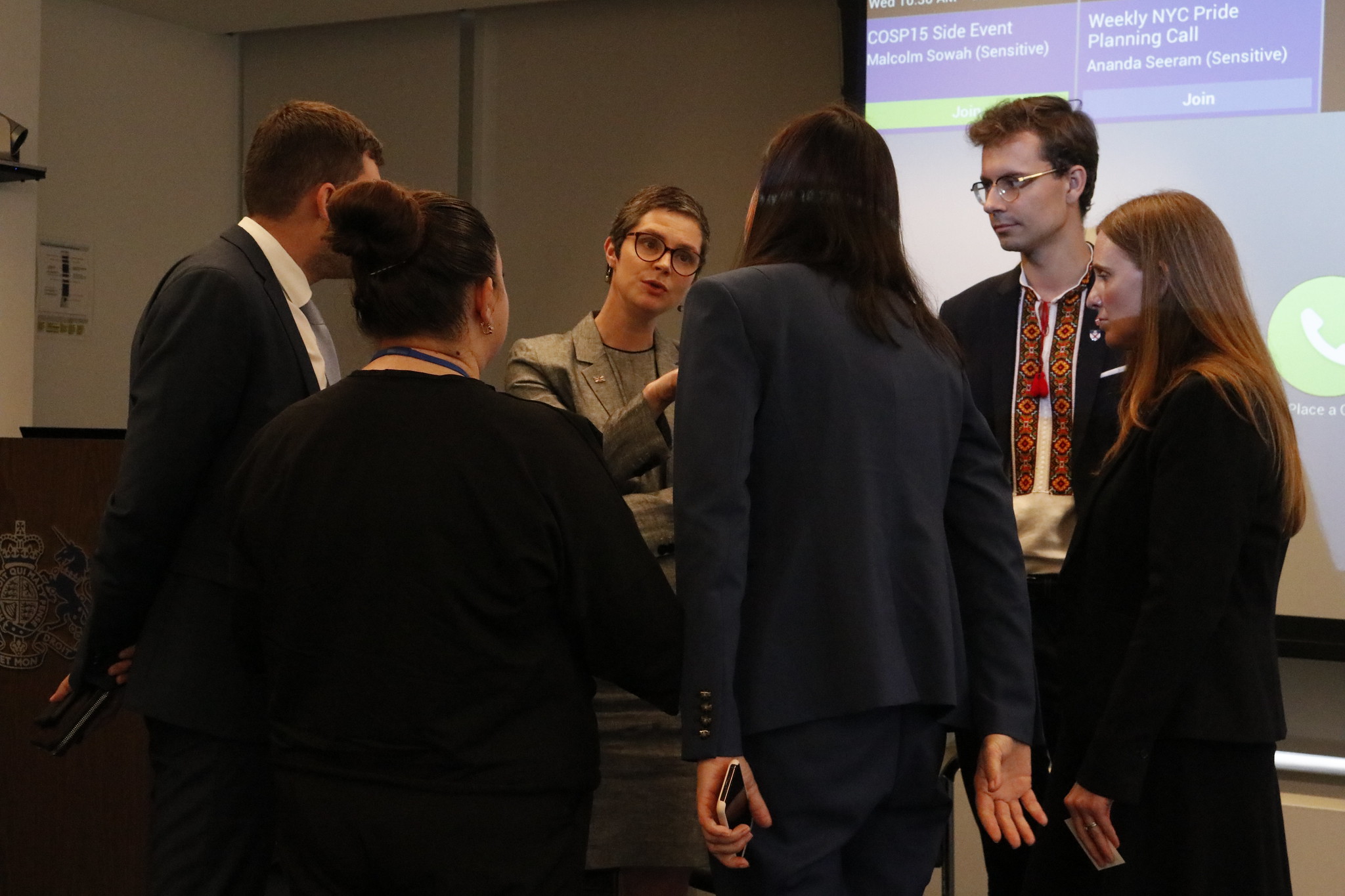 The Minister for Disabled People faces forward speaking with a small group of members of the Ukrainian delegation. The delegation have their backs or side to the camera facing towards the Minister wearing dark formal clothing. One male member has a traditional tie type scarf which is red with circular motives and a red tassle.