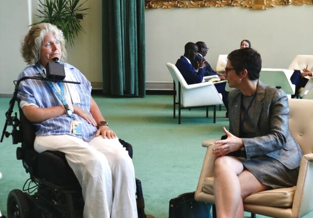 The Minister for Disabled People is sat down with her legs crossed in a grey suit and skirt, she is facing and talking with Rosemary Kayess, Chair of the UNCRPD Committee. Rosemary is wearing white trousers and a blue top and is sat in a motorised wheelchair with her arm in a white sling and a chin support support her head.
