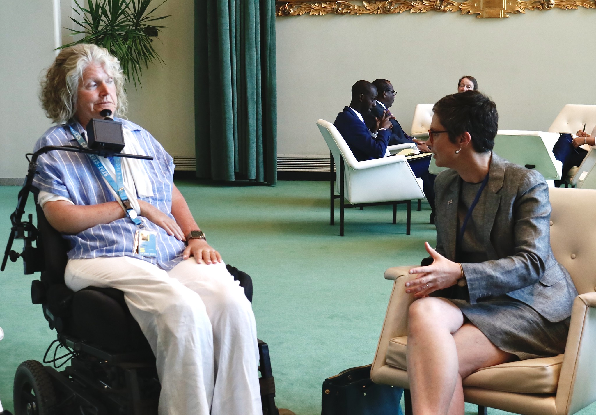 The Minister for Disabled People is sat in a green carpeted room with her legs crossed in a grey suit and skirt, she is facing and talking with Rosemary Kayess, Chair of the UNCRPD Committee. Rosemary is wearing white trousers and a blue top and is sat in a motorised wheelchair with her arm in a white sling and a chin support support her head.