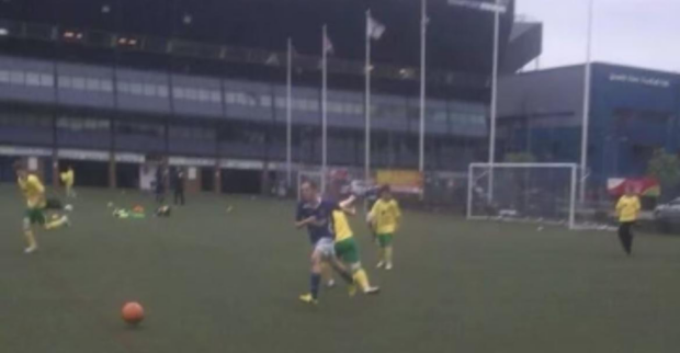 An action shot image of Harry in a blue shirt in the centre of the image chasing an orange football on a football field. 