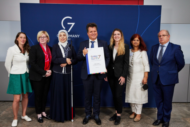G7 delegates standing in front of a G7 Germany sign. Commissioner Dusel is holding a large copy of the Chair’s Summary.