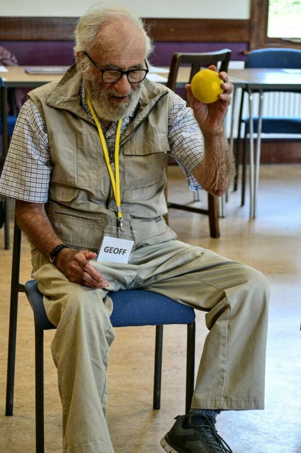 A older chap with black rimmed glasses sat down. He is looking downwards, holding a yellow ball in his left hand ready to throw it. He is wearing light coloured trousers, waistcoat and shirt.