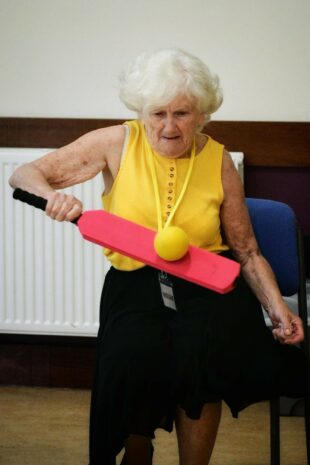 A older lady with white hair wearing yellow sleeveless top and black skirt. She is standing holding a red bat in her right hand hitting a yellow ball. 