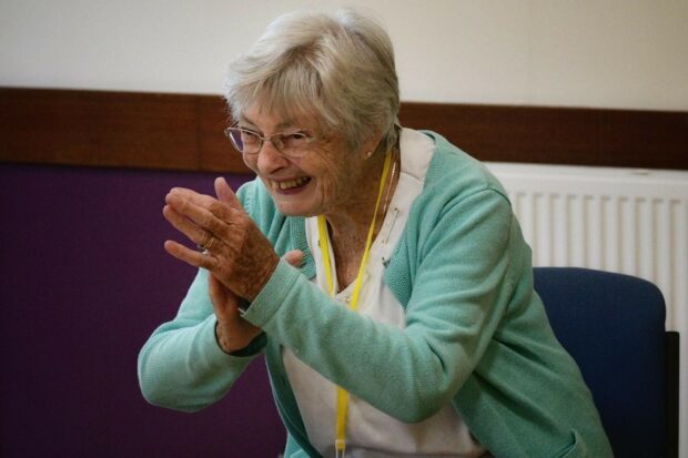 Older lady leaning forward off a chair with her hands clapped together. She is wearing glasses, a light turquoise cardigan and glasses. 