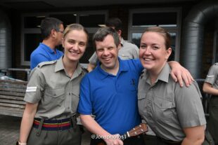 Two female Royal Marines wear their khaki uniforms with a male participant of the project wearing a blue t-shirt stood in the middle of them. All are smiling and looking at the camera.
