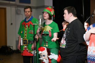 From left to right a male singer wearing an elf t-shirt looking to the middle, a female singer wearing an elf hat with ears and an elf t-shirt singing into a stand up microphone with her hand outstretched, and a male singer wearing a black festive t-shirt looking towards the microphone. 