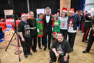 Royal Marine musician in red-breasted tuxedo with medals and a colleague in regimental outfit stands with participants of the project either side. The participants are wearing Christmas themed t-shirts, Christmas jumpers, elf outfits and Father Christmas hats.