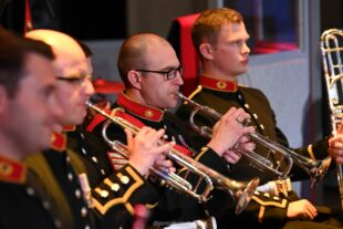 Four Royal Marine musicians sat in a line wearing their full dress uniform with red collars, black jacket and gold buttons. Two are holding up trumpets playing and one holds a trombone. 