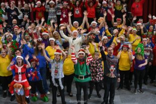 Alt text: Group of people cheering with their hands up all wearing Christmas jumpers and bright outfits of yellows, blues and purples. Some people are wearing Christmas jumpers, tinsel or elf outfits. In the middle a chap wears a white tuxedo suit. 