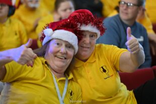 Two female members of the Music Man Project wearing yellow polo t-shirts with the Music Man Project logo on it of a person wearing headphones. They are both wearing Christmas hats with tinsel, hugging each other, smiling and holding their hands up in a thumbs up gesture.